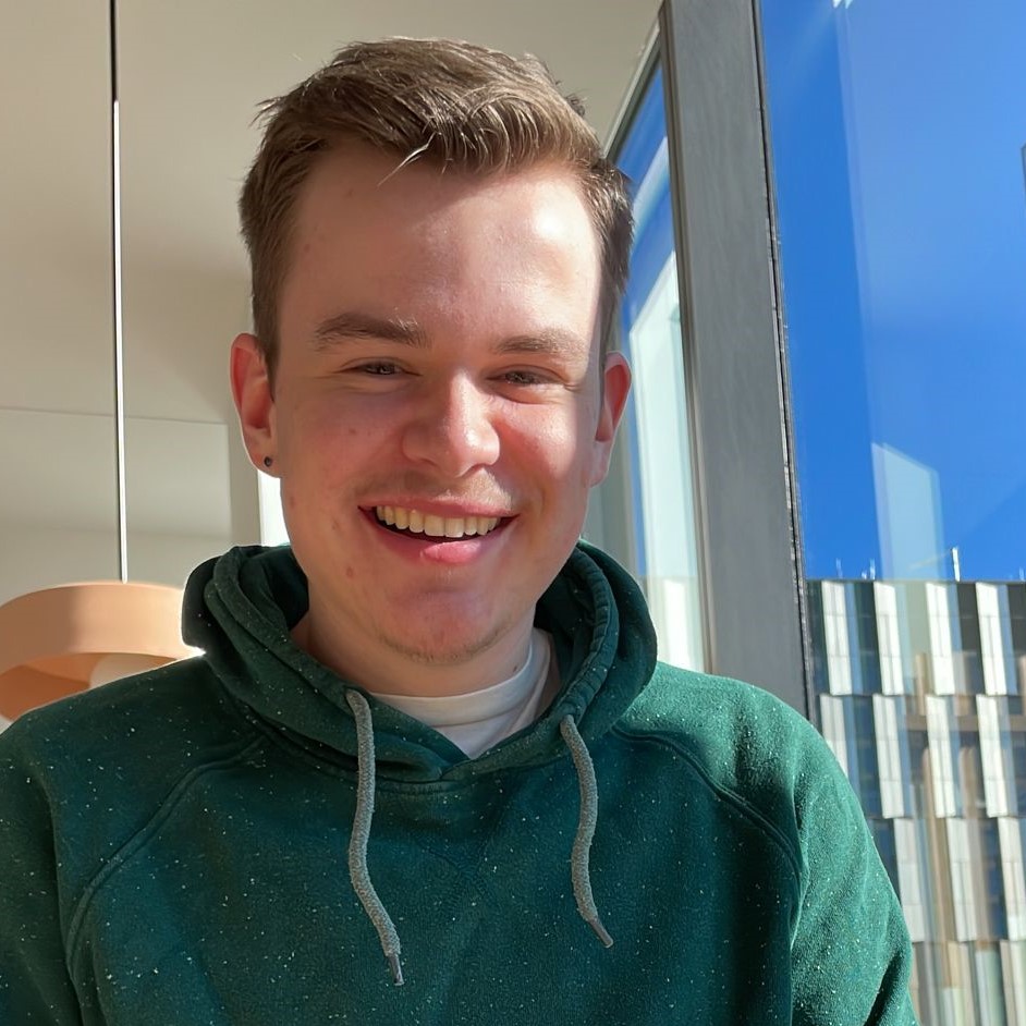 head shot of Thomas, a man with short blond hair in green sweater in front of a window